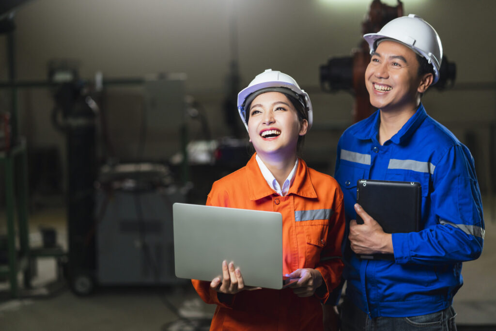 portrait of asian emgineer male female technician in safty uniform standing and turn around to look at camera and laugh smile with cheerful and confident in machinery factory workplace background