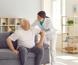 Female nurse assisting happy senior patient with daily activities in nursing home. Young woman helping aged man to stand up from couch in room of modern hospital or geriatric rehabilitation center